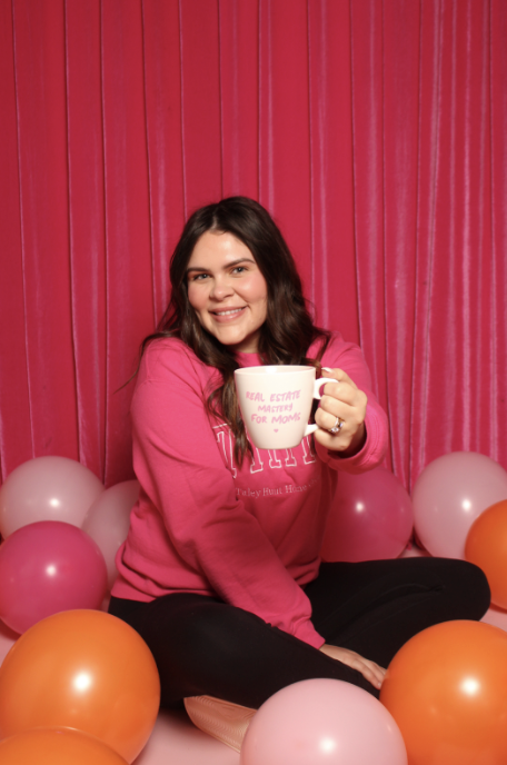 Taley Hunt sitting in a pink room with balloons, holding a mug that says “Real Estate Mastery for Moms."